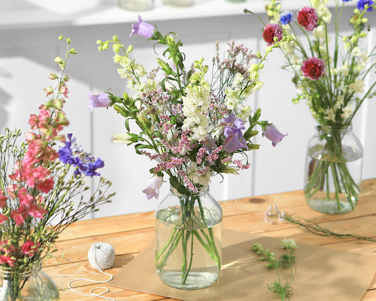 Meadowy bouquets in glass vases are lined across a wooden table. A few stems and a ball of twine lie between them.