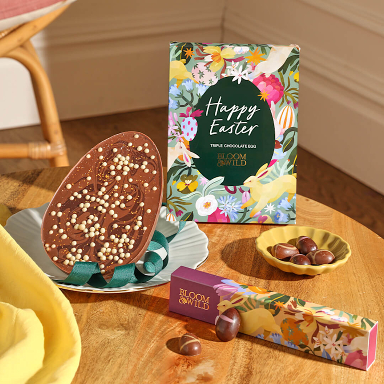 A flat, triple-chocolate Easter egg decorated with crispy white chocolate pearls, paired with a floral "Happy Easter" gift box and chocolate truffles.