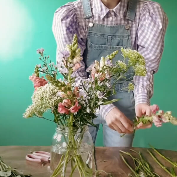 The glass vase is now filling up with more stems at various heights. Harriet holds another stem, about to place it in.