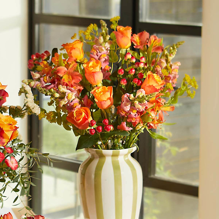 A vibrant bouquet of apricot orange roses, delicate pink snapdragons, and alstroemeria with deep red berries and yellow solidago, arranged in a white vase with green vertical stripes.