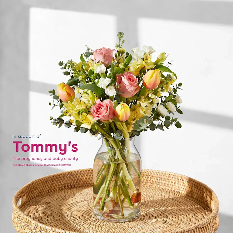 A close-up of a bouquet with a charity message overlaid. The text reads "In support of Tommy's The pregnancy and baby charity Registered charity number 1060508 and SC039280". A purple tulip and a white iris are prominent in the foreground.