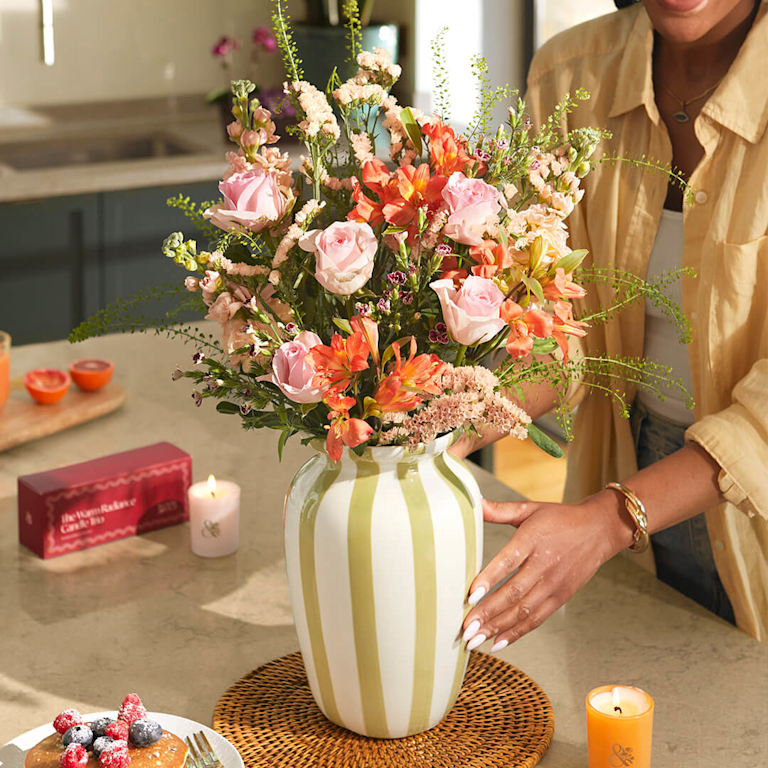 A warm, joyful bouquet featuring soft pink roses, orange alstroemeria and delicate lisianthus in a green-striped vase. The arrangement offers a mix of velvety petals, creating a bright and inviting kitchen atmosphere.