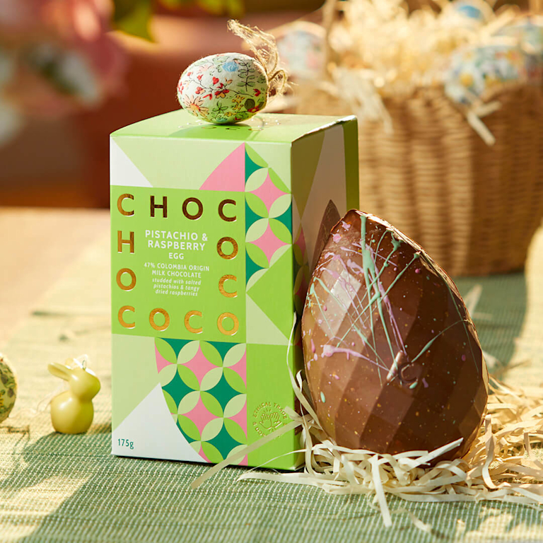 A milk chocolate Easter egg speckled with green pistachio and pink raspberry, beside a vibrant green gift box.