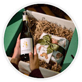 Hands opening a green gift box with a bottle of wine and festive food treats nestled in straw.