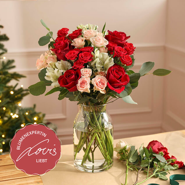 Festlicher Fairtrade-Strauß mit tiefroten und zartrosa Rosen sowie weißen Alstroemerien in einer Glasvase, mit Eukalyptuszweigen und Weihnachtsdeko im Hintergrund.