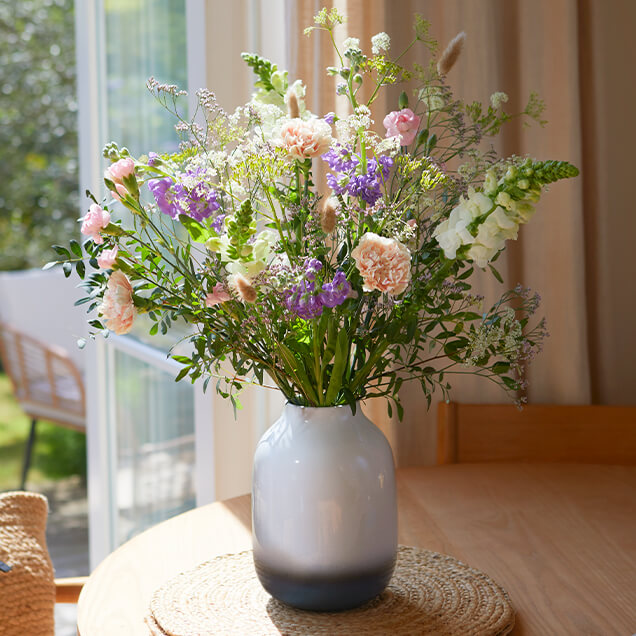 Ein Blumenstrauß in zarten Farbtönen steht in einer Keramikvase neben dem Fenster