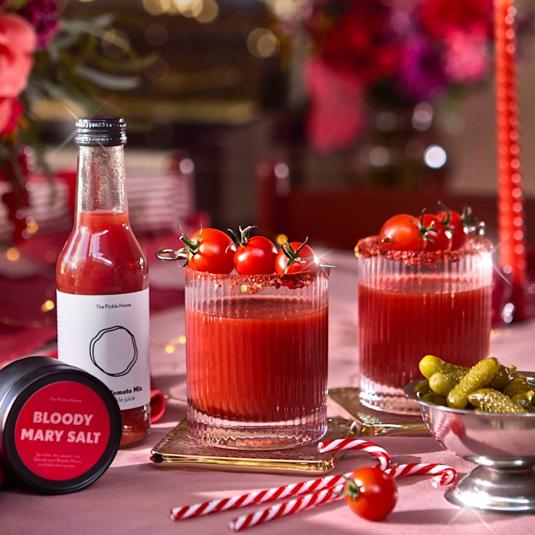 Two Bloody Mary cocktails garnished with cherry tomatoes, beside a bottle of Bloody Mary mix, a tin of Bloody Mary salt, gherkins and candy-cane straws.
