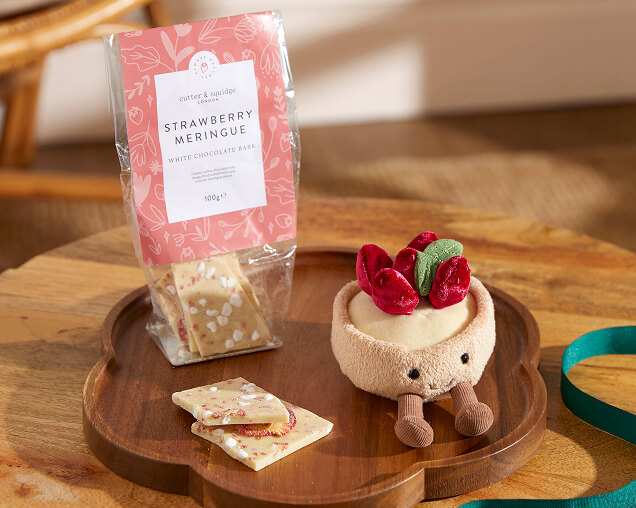 A pouch of strawberry meringue white chocolate bark next to a round tan plushie of a cherry tart on a wooden tray.