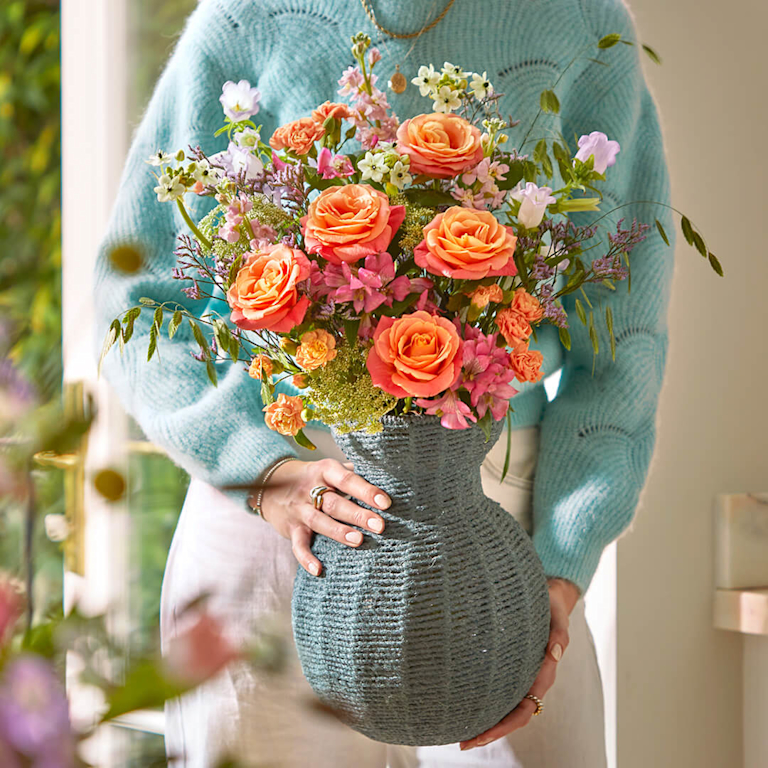 A vibrant bouquet of coral roses and apricot spray roses, accented with lilac campanula and delicate white lace flowers. It's held in a textured, slate-blue woven vase.