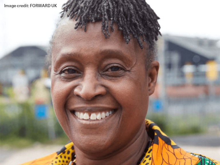 Naana, a dark-skinned older woman with tightly coiled haired, wears a shirt in a traditional Ghanian print and smiles broadly.