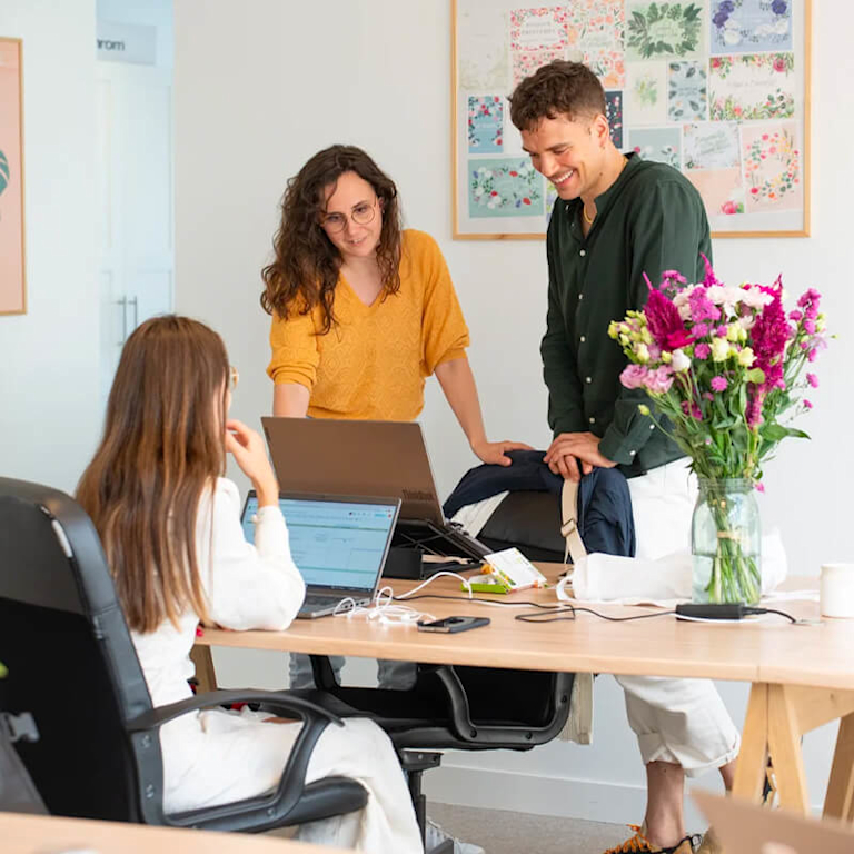 Two women and a man father around a desk in the light, airy Bergamotte office. A vase of flowers is on the table.