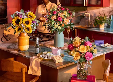 Three bouquets on a marble kitchen island: sunflowers in a yellow vase, pink and peach roses in a sage vase, and pink and yellow roses in a glass jug, with prosecco, chocolates and gifts in warm light.