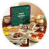 Birthday gift box on a red gingham cloth, surrounded by scones, jam, chocolate bars, mini cakes and a card reading "Happy Birthday to you".
