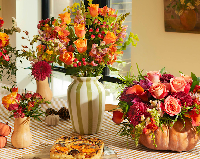 Three autumnal flower bouquets and a pumpkin vase. The largest is a bouquet of orange, red, and yellow roses in a cream vase with green stripes. It's sat on a plaid tablecloth next to a focaccia.