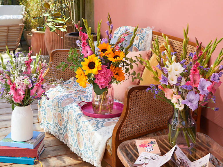 A sunny outdoor scene featuring three vibrant bouquets. One bouquet, with sunflowers and pink hydrangeas, sits on a wooden bench with patterned cushions. Two other bouquets, bursting with colourful gladioli and stocks, stand nearby.