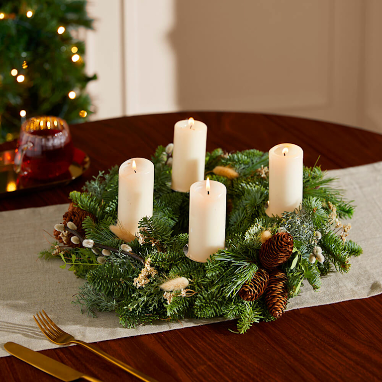 Traditioneller Adventskranz aus Tannengrün, verziert mit getrockneten Blüten, Weidenkätzchen und Tannenzapfen, mit vier brennenden, cremefarbenen Stumpenkerzen. Er liegt auf einer Holzoberfläche mit hellem Tischläufer und goldenem Besteck.