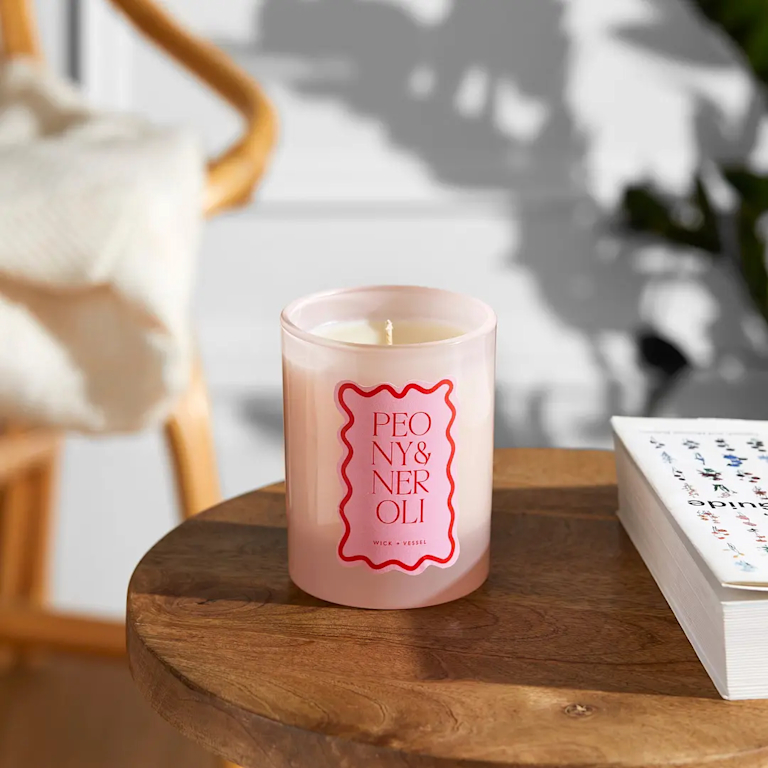 A candle sits on a round wooden table. The candle has a white label outlined in red with the text PEONY & NEROLI stacked vertically in red lettering. The table also holds a book with a partially visible spine.