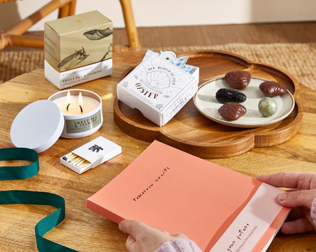 A top-down view of a wooden table featuring a lit "Sweet Pea & Jasmine" candle, a peach-colored notebook held by two hands, a small box of matches, and shell-shaped chocolates on a wooden tray.