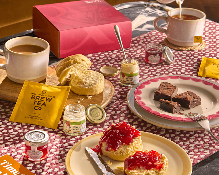 A wicker gift hamper filled with birthday treats including vanilla fudge, shortbread, honeycomb, NIO cocktails, and a "Birthday Cake" gift card, with a hand reaching in and a decorated cake slice visible.