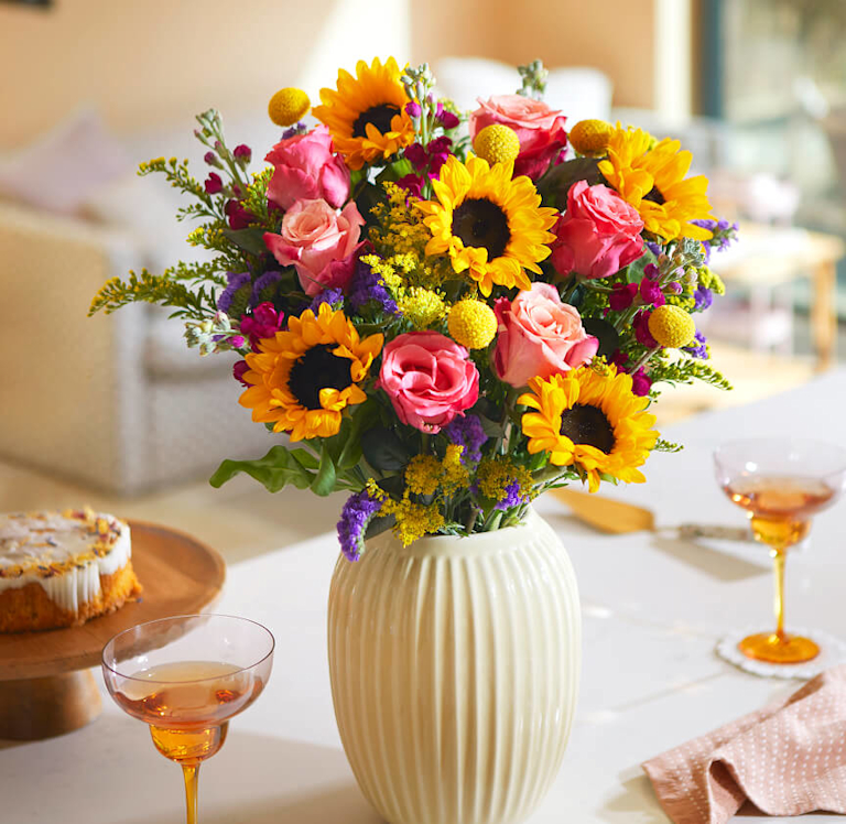 A radiant bouquet of sunflowers, pink roses, and purple statice in a cream ribbed vase. Yellow craspedia and solidago add texture. The arrangement sits on a white table with two amber-coloured glasses and a cake.