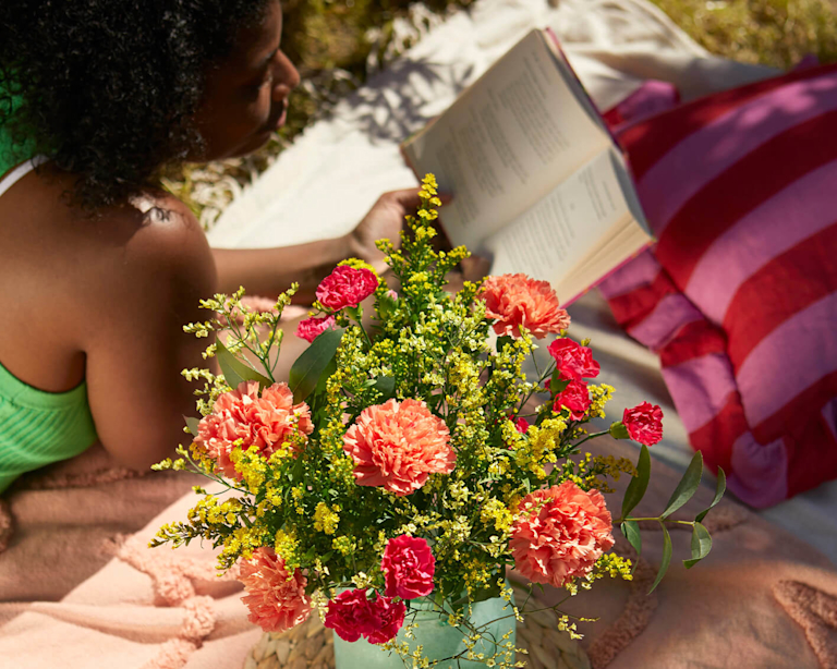 A dark-skinned woman with curly hair lies on a beach towel and reads a book. A summery bouquet of yellow and green stems sits next to her.