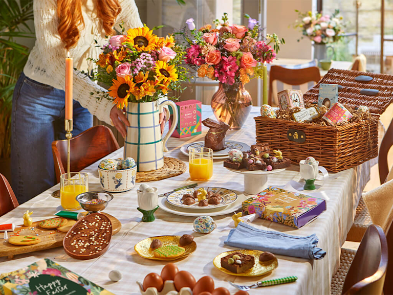 A sun-drenched Easter table featuring vibrant sunflower and pink rose bouquets, a wicker hamper filled with chocolate treats, and fudgy brownies. The scene is full of spring joy, combining fresh florals with sweet bakes.