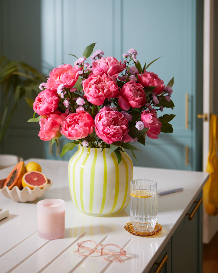 A vibrant bouquet of large, ruffled, coral charm peonies and delicate sprigs of lilac flowers, arranged in a modern, white vase with bright lime green vertical stripes. The overall impression is cheerful and summery.