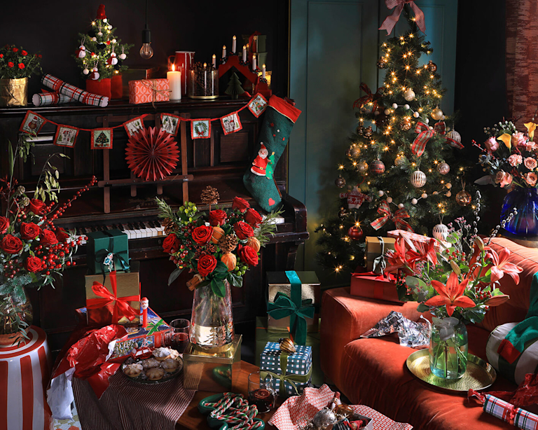 An abundant Christmas scene. Festive-coloured flowers, wrapped presents, sweet treats and Christmas crackers are spread across the room. A Christmas tree stands in the right corner.