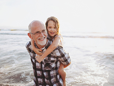 Elderly man piggy-back carrying his grandchild by the water shore