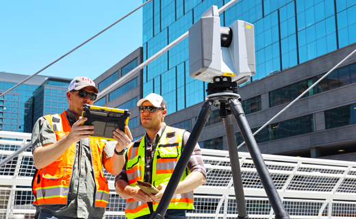 Two surveyors, one holding a Trimble tablet, the other looking at its screen, next to a Trimble X12 laser scanner in city setting.