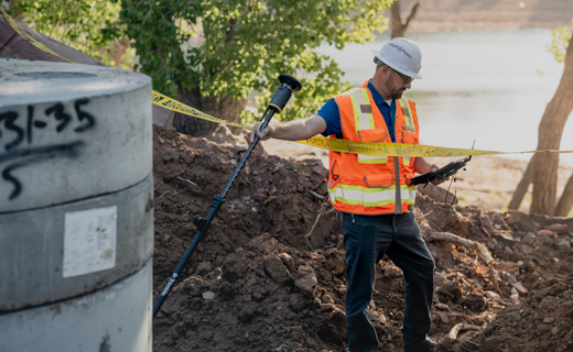 A surveyor in a hard hat and safety vest stands in a dirt trench beside a concrete pipe, holding a tilted ST30 smart target in one hand and a surveyor controller in the other.