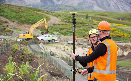 Two construction workers on site reviewing data on a Trimble TSC6 with a digger and truck in background