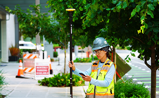 Surveyor standing in city street looking at smartphone in her hand while holding a survey pole with Trimble DA2 receiver on top with the other hand.