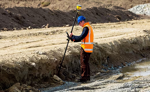 Géomètre sur un chantier près d'un talus, tenant une canne de levé inclinée et surmontée d'un récepteur Trimble R780.