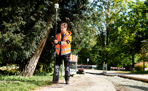 Surveyor holding a survey pole with Trimble R12i receiver on top; man wearing high-visibility clothing standing on sidewalk next to some trees. 