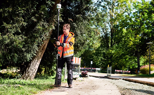 Géomètre tenant une canne de levé surmontée d'un récepteur Trimble R12i ; homme portant des vêtements à haute visibilité se tenant sur le trottoir près de quelques arbres.