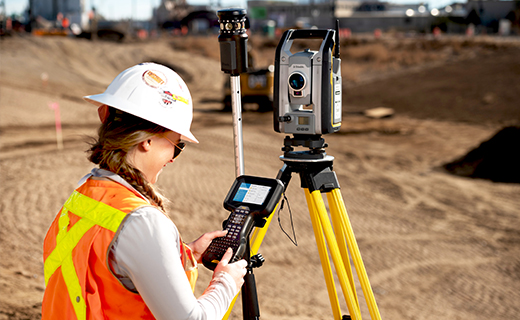 Surveyor standing on a construction site holding a Trimble TSC5 survey controller in her hands.