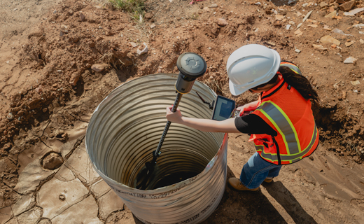 A contractor in a hard hat and safety vest stands over a large metal culvert pipe embedded in the dirt. She is holding the ST30 smart target inside the culvert and looking at her survey controller.