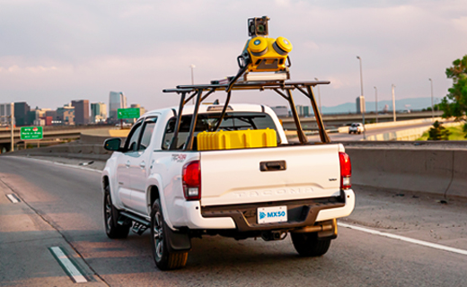 Trimble MX50 mobile mapping system mounted on top of a white SUV vehicle which is driving on a freeway towards high rise city buildings.