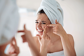 Young woman in mirror squeezing pimple on face