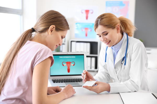 W gabinecie lekarka rozmawia z młodą kobietą, pokazując jej coś na niewielkim kartoniku. W tle widać laptop i plansze.
