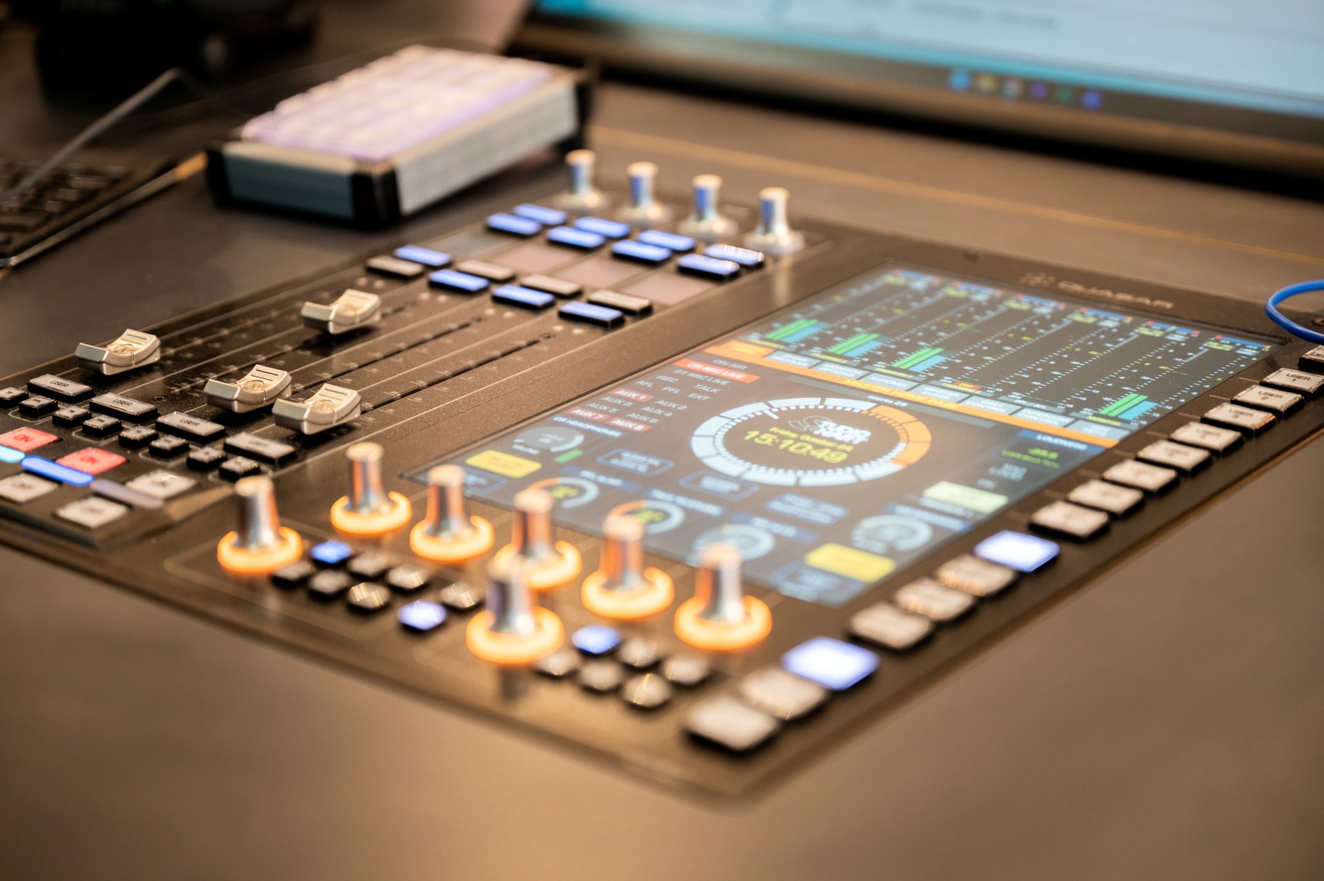 Close-up van een audio mengpaneel met verlichte draaiknoppen, faders en een digitaal touchscreen waarop de geluidsniveaus en instellingen worden weergegeven.