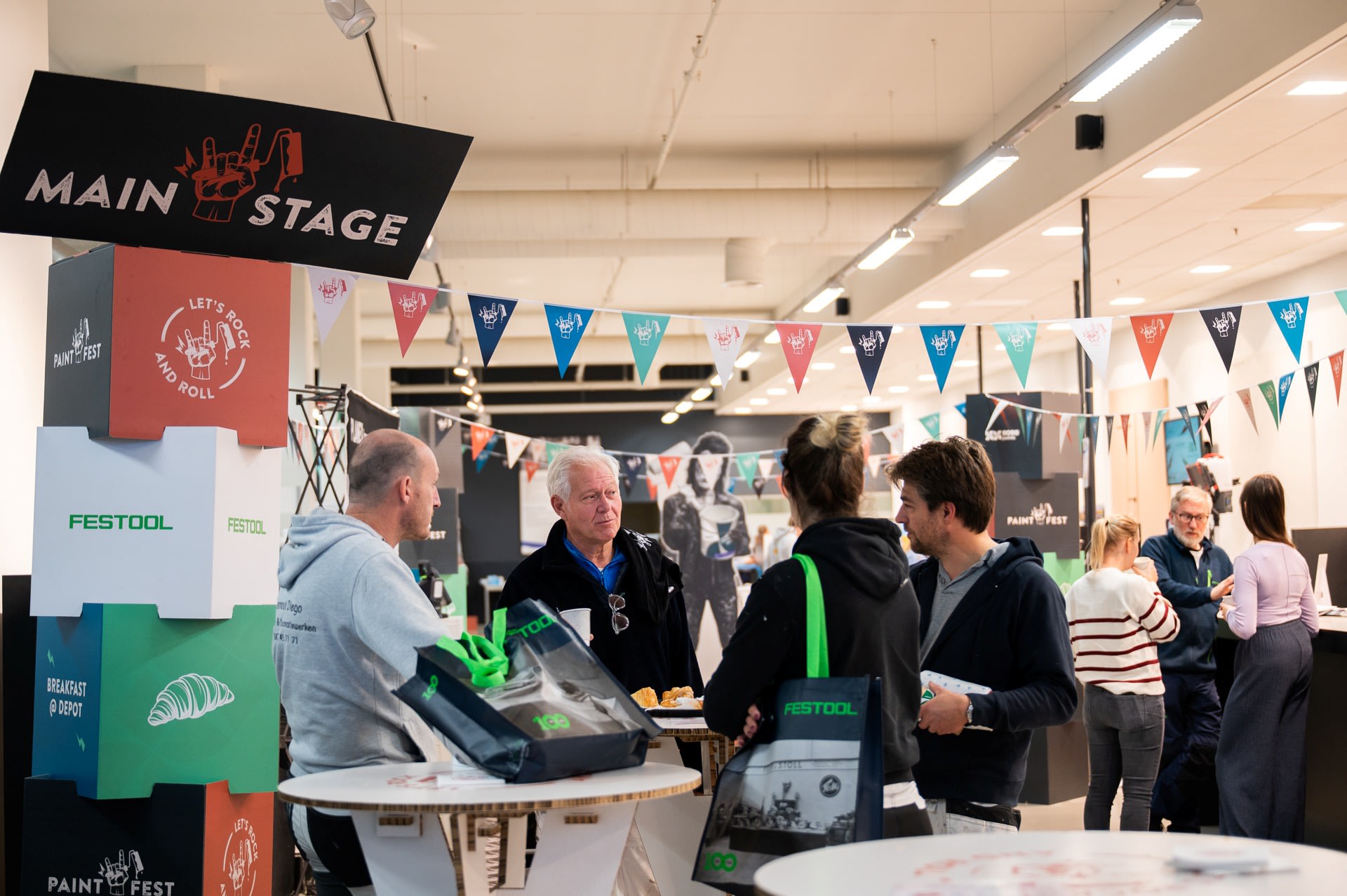 Mensen komen samen en praten op een indoor evenement of expo, met merkstands, vlaggen en informatieve displays zichtbaar op de achtergrond.