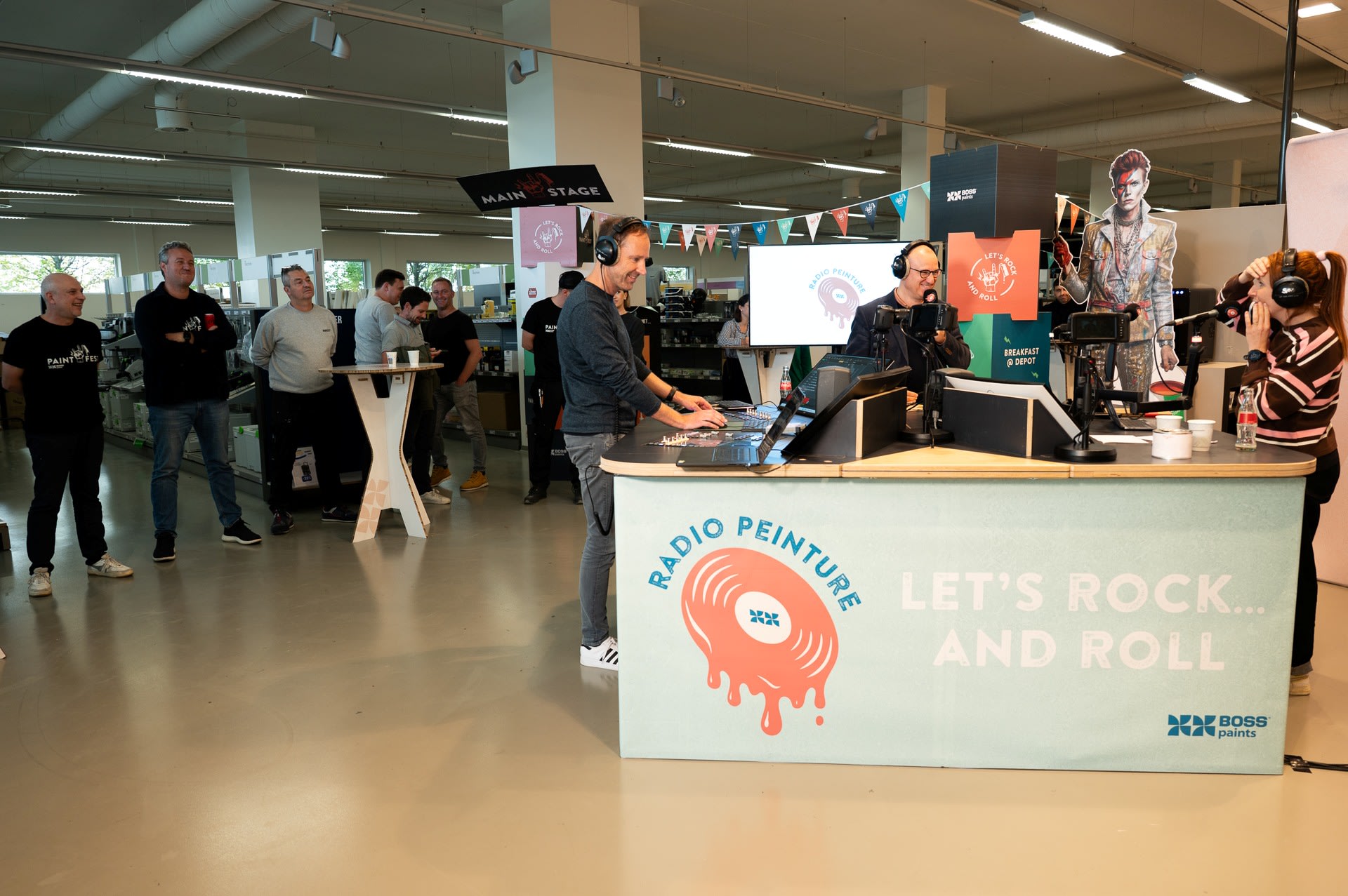 Mensen hosten een live radio-uitzending in een versierde cabine in een modern gebouw terwijl anderen in de buurt toekijken.