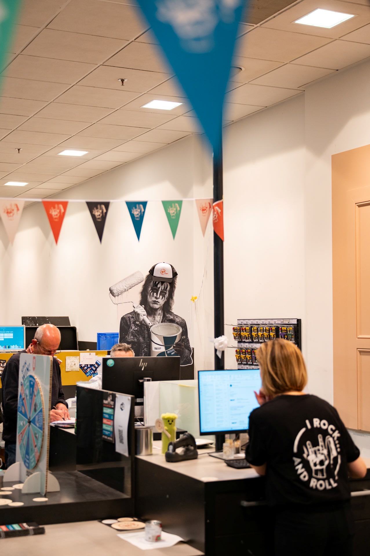 Mensen werken aan bureaus in een kantoor versierd met kleurrijke driehoekige spandoeken; op de achtergrond is een kartonnen knipsel te zien van een persoon die een verfroller vasthoudt.