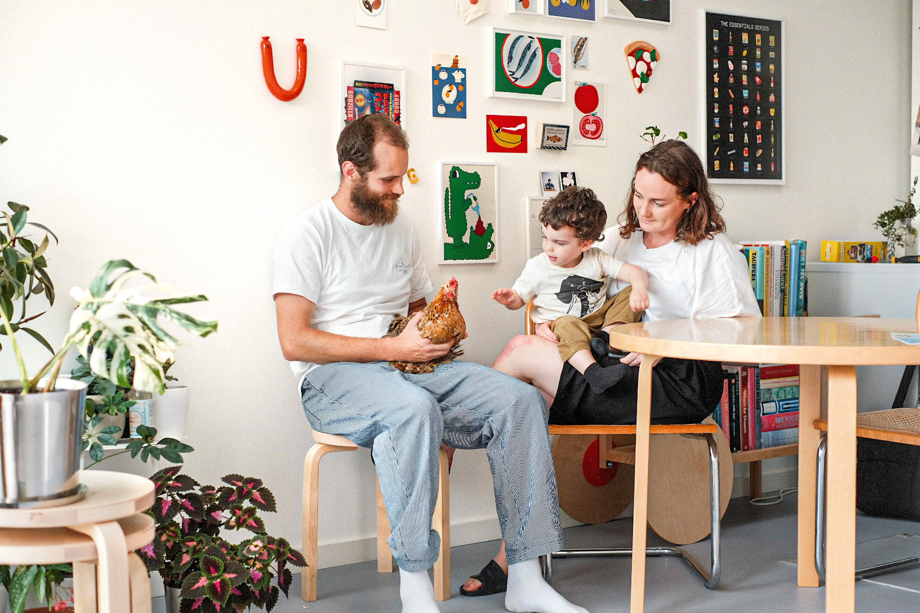 Een volwassene en een kind zitten met een andere volwassene die een kip vasthoudt in een vrolijk ingerichte kamer met planten, kunst en boekenplanken.