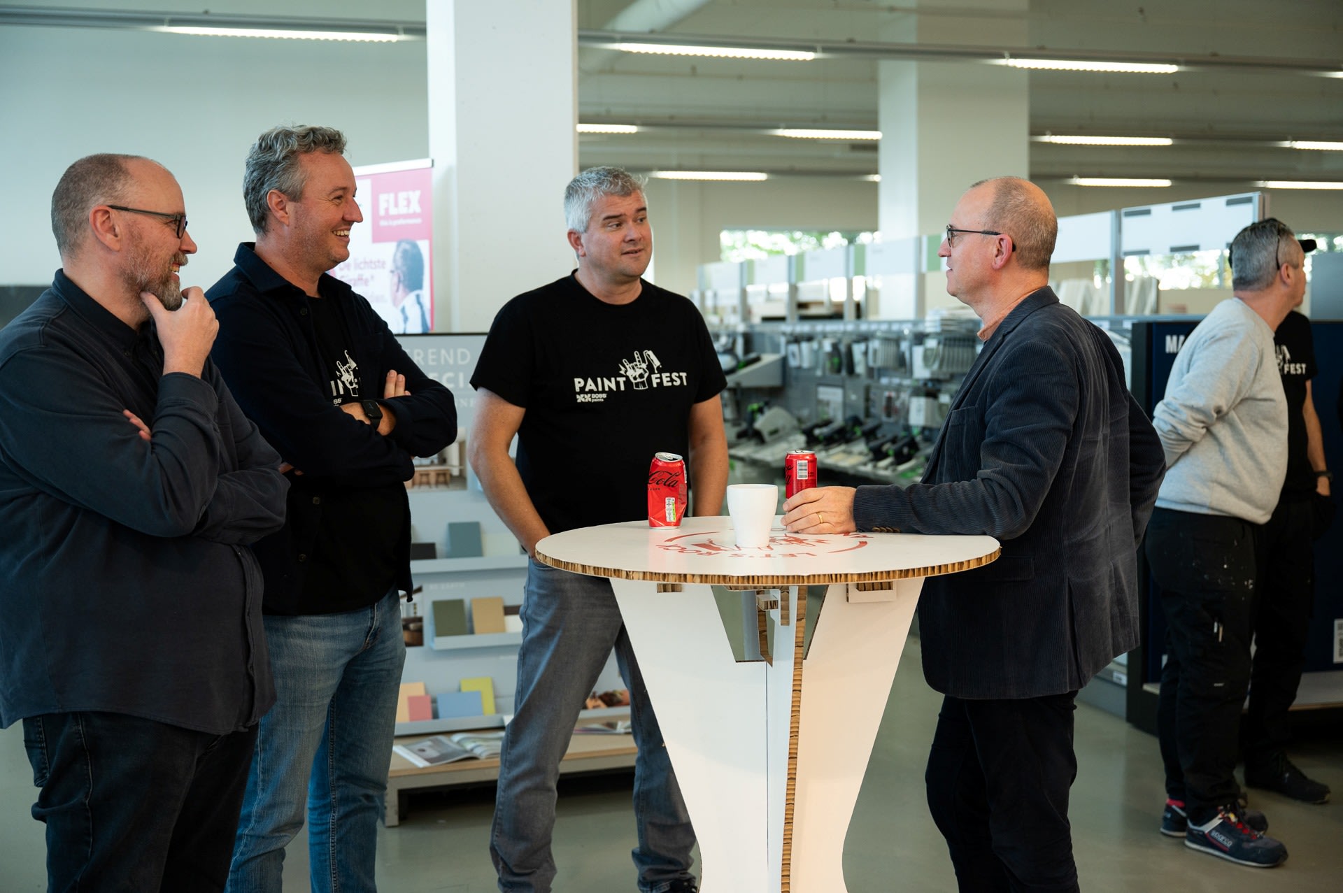 Vier mannen staan te praten rond een hoge tafel met drankjes in een helder verlichte omgeving; een andere man staat op de achtergrond bij planken.
