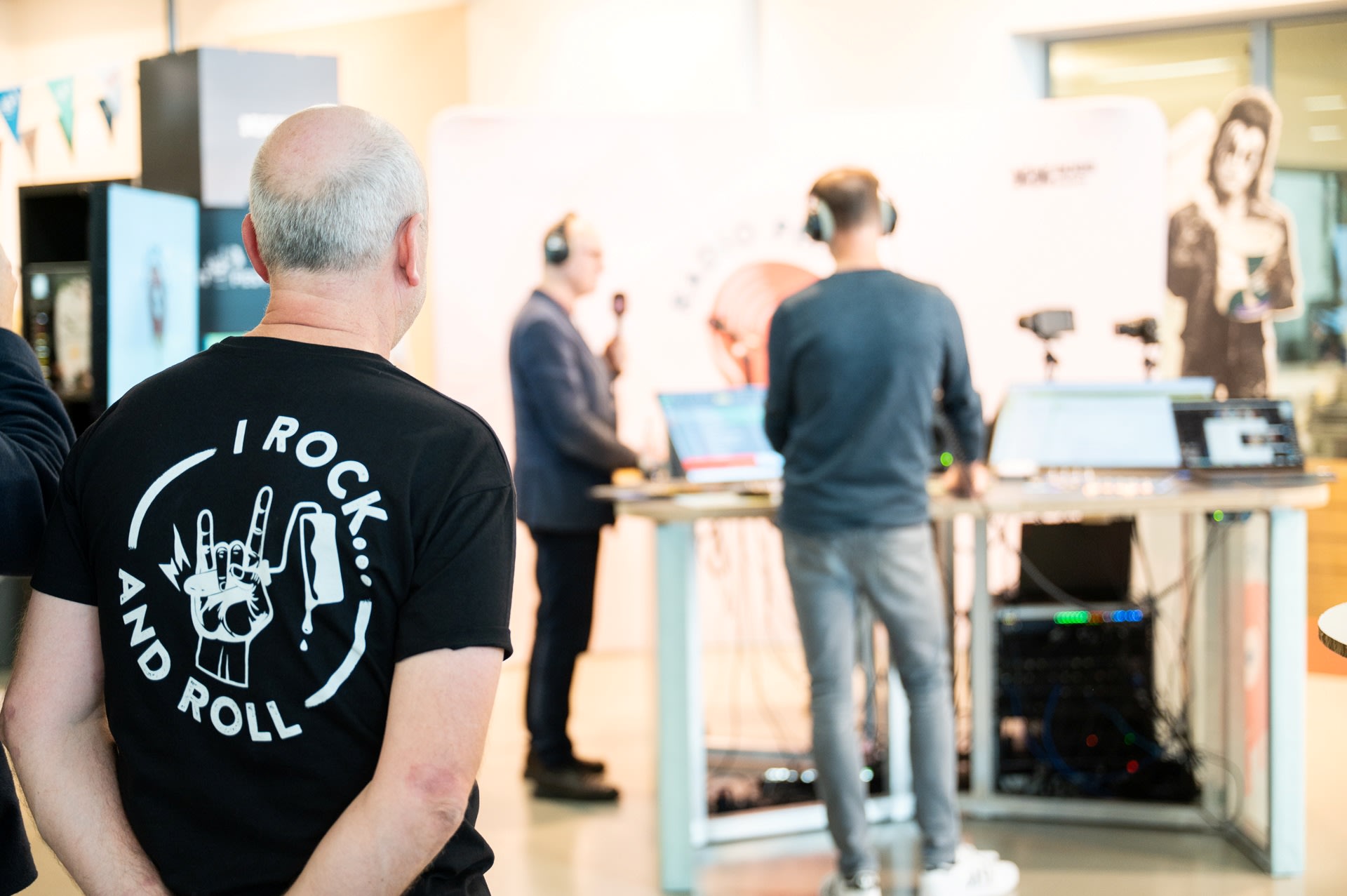 Een persoon met een shirt met de tekst "I rock and roll" staat op de voorgrond, terwijl twee mensen met koptelefoons op op de achtergrond een uitzending volgen.