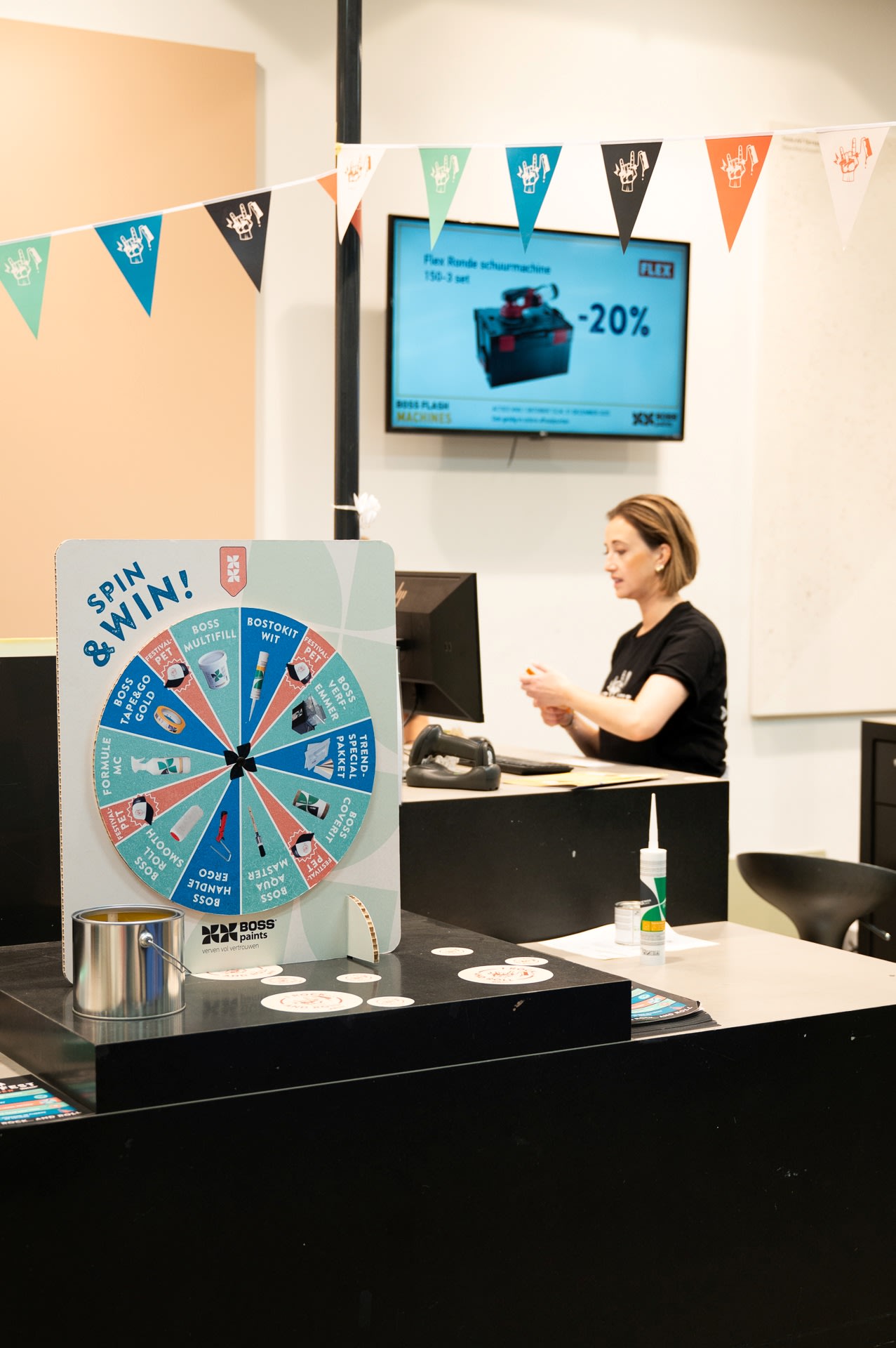 Een vrouw zit aan een bureau achter een toonbank met een "Spin & Win!" prijzenwiel; op de achtergrond hangt een promotiescherm aan de muur.