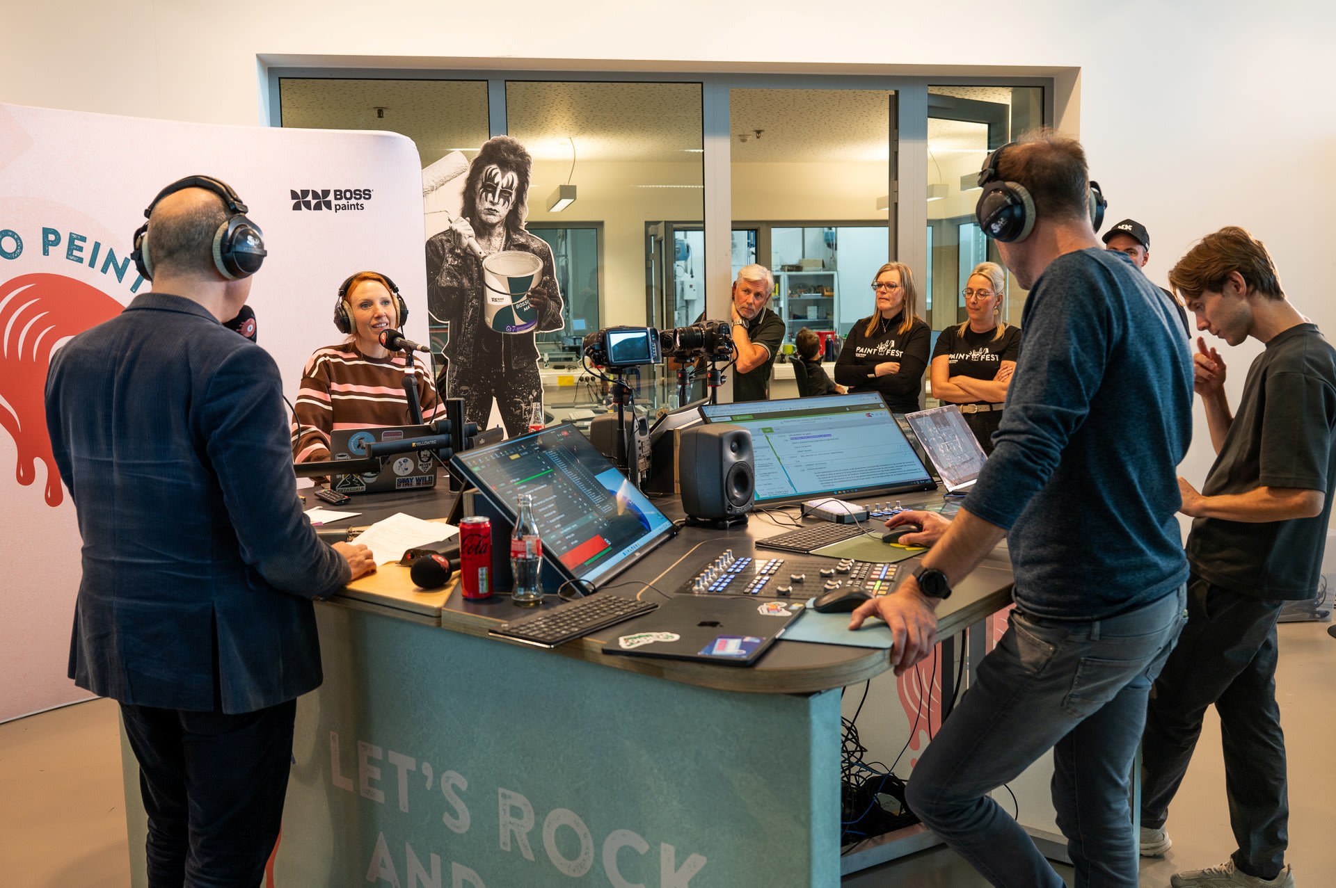 Radiopresentatoren zitten aan een studio met microfoons en koptelefoons, terwijl verschillende mensen door een raam op de achtergrond staan te kijken.